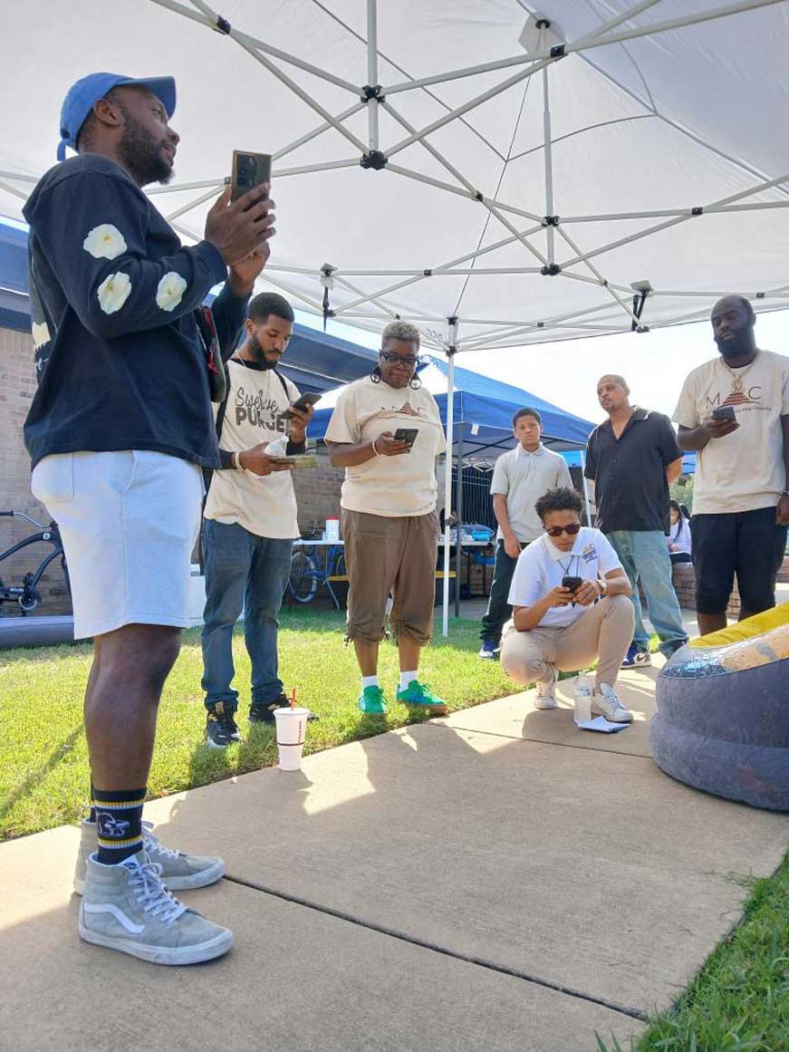 A group of people stand and gather under a white canopy outdoors, looking at their phones or listening attentively. Several wear beige ‘Memphis Artists for Change’ shirts, while one person in front crouches down holding a phone. The setting appears to be a community event on a sunny day near a brick building.