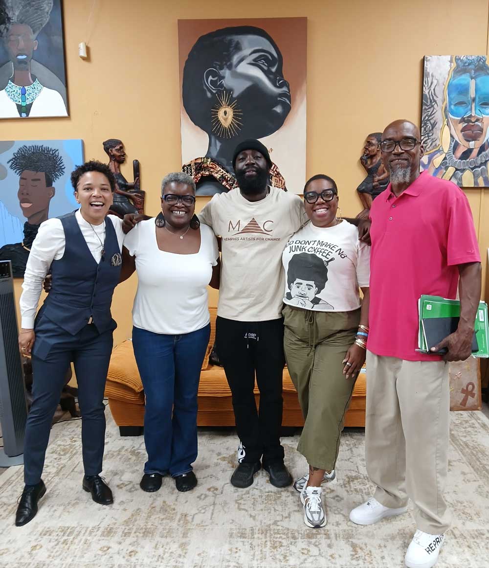 Five people stand together smiling in front of a wall decorated with portraits of Black women in Afrocentric art styles. The group includes three women and two men standing arm in arm. One person wears a shirt that says ‘Memphis Artists for Change,’ and another wears a shirt that reads ‘God Don’t Make No Junk Cheef.’ The setting appears warm and creative, highlighting community and collaboration.