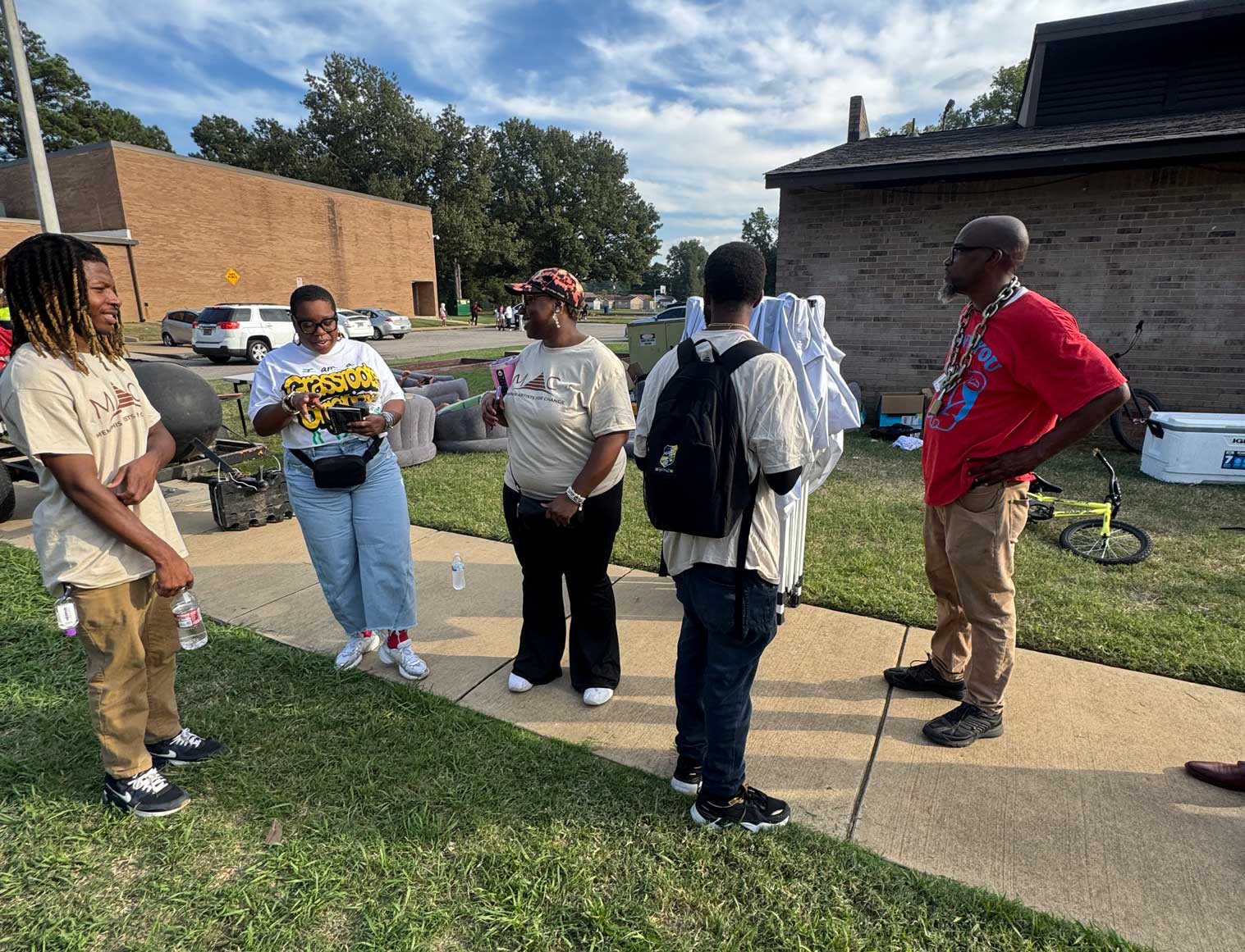 A group of five people stand outside on a sunny day having a conversation. Three wear matching tan shirts with a logo, while one person in a white shirt and jeans holds a yellow sign that says ‘Grassroot.’ Another person in a red shirt stands to the right with hands on hips. They are gathered on a sidewalk near a grassy area, with a brick building and parked cars in the background.