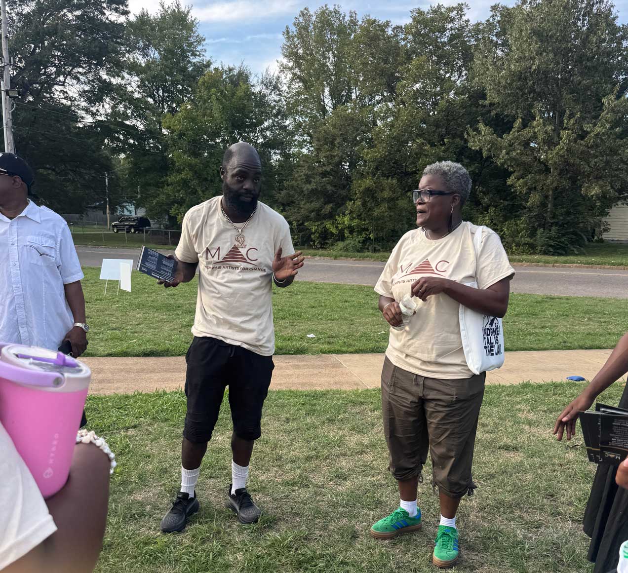 Two people wearing matching beige ‘Memphis Artists for Change’ T-shirts stand outdoors on a grassy area having a conversation. The man holds a tablet while gesturing as he speaks, and the woman listens, holding items in her hands. Trees and a residential street are visible in the background, suggesting a community event.